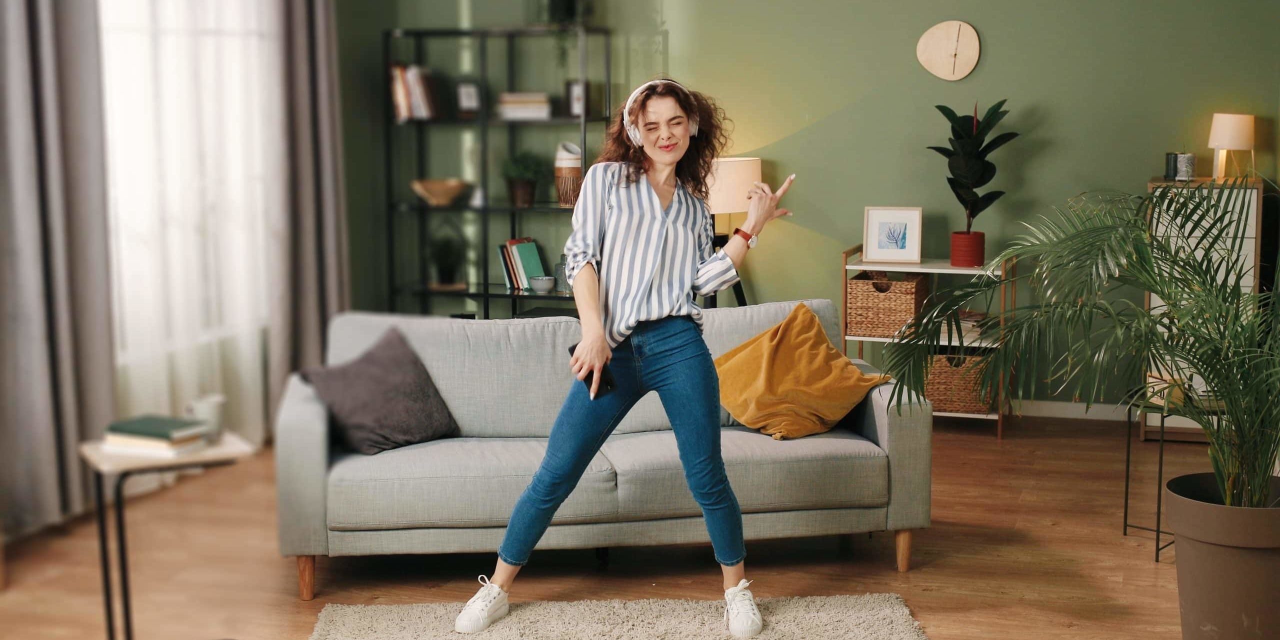 Happy joyful young Caucasian beautiful woman in positive mood dancing moving rhythmically and jumping while listening to music song in headphones on smartphone, having fun in room, leisure concept